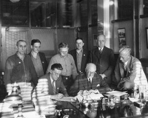 Harold Christoffel (seated, left) and Harold Story (seated, right) sign a contract surrounded by fellow Allis-Chalmers employees and managerial representatives. 