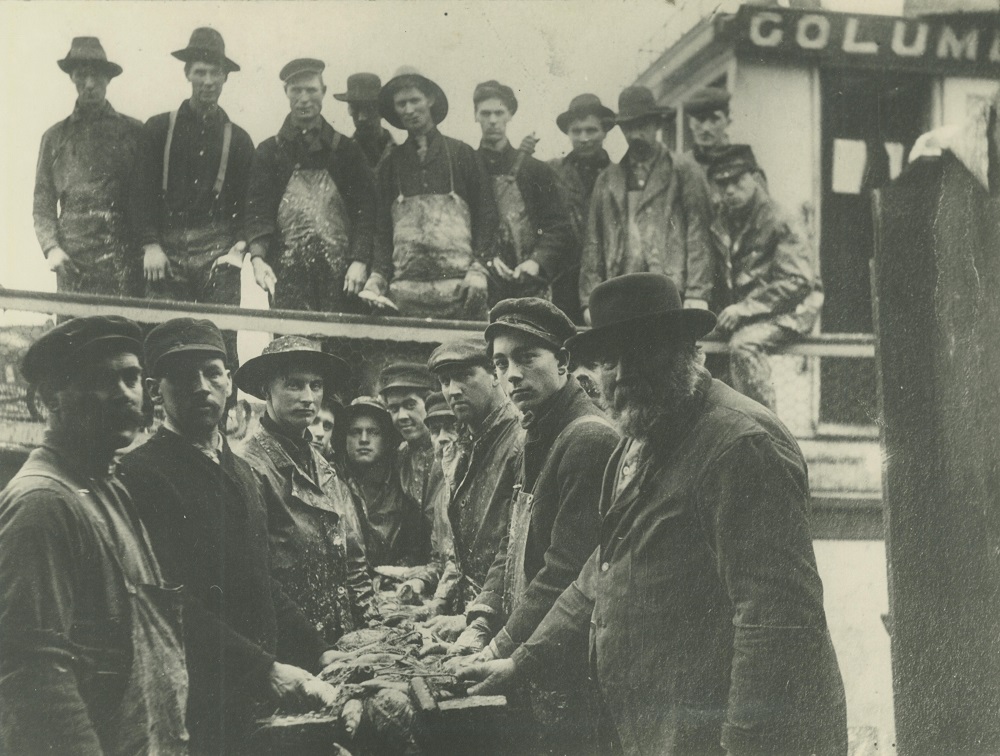 A group of fishermen pose with with their catch. The men in the background stand on a fishing boat while the men in the foreground hold nets. 