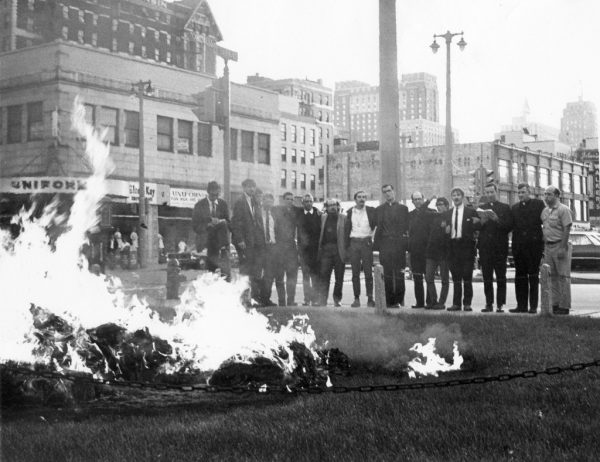 Photograph of the fourteen men who burned approximately 10,000 draft cards in 1968 standing arm-in-arm. The man furthest to the left is a newspaper reporter. 